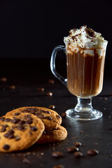 Close up of a glass cup of coffee with whipped cream and chocolate on it, chocolate chip cookies and roasted coffee beans on dark background. Concept of ready to eat food, tasty snack. Selective focus