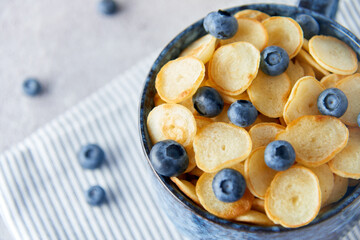 Blue Bowl of mini pancake cereal with blueberries, maple syrup and milk on concrete background. Tiny pancakes, new food trend concept. Cute healthy breakfast or snack. Recipe, menu.