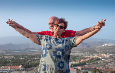 Mature couple of married people standing on hill with mountain and city on background looking at camera with arms raised - active seniors and freedom concept