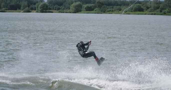 Wakeboarding in a lake near the forest, sportsman surfs on the water, ride on a wakeboarding board, performs tricks and does a somersault, 4k slow motion.