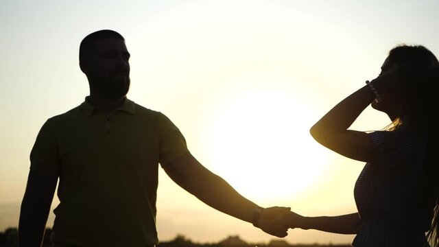 Happy young guy and girl enjoy romantic evening on nature in field holding hands and dancing against backdrop of summer sunset close-up view in slow motion. Date of couple in love outdoors in country.