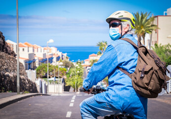 Fototapeta na wymiar Retired senior man in deserted road with bicycle and yellow helmet wearing face mask due to coronavirus. Blue sky and horizon over the sea.