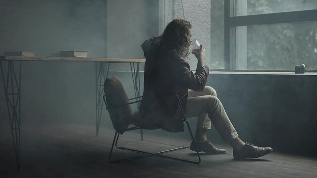 handsome and charismatic guy with long curly hair and beard, in a brown leather jacket, light pants and shirt, sitting on a chair in loft studio, holding a whiskey, moving with music and enjoying life