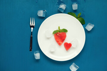 White plate with cut out watermelon hearts, mint and ice on blue wooden background. Flat lay.