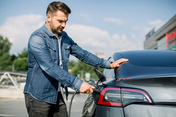 Side view of young handsome bearded man in jeans shirt, plugging wire into the car socket to charge...