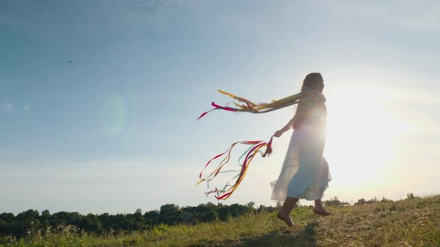 A teenage girl in a white dress is having fun, runs barefoot on the top of a hill with rainbow ribbons in her hands against the background of the sky and the sunbeams