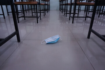 The used medical face mask is placed on the classroom floor with wood lecture chairs in the empty classroom. Concept during the Coronavirus disease COVID-19 in the 2020s. Back to school concept.