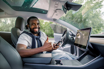 Cheerful smiling African man driver sitting in his new high tech electric car with upgraded avtopilot self-driving system, holding his smartphone and showing thumb up