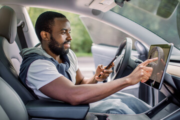 Young excited African man working with car touchscreen display and mobile car app, while sitting in...