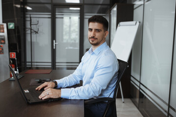 dark-haired man in the office in a blue shirt sits at a table in front of a laptop, putting his hands on the keyboard