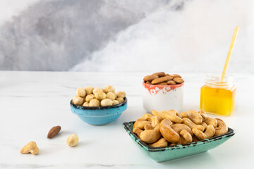 Beautiful bowls of different colors with roasted nuts (cashews, almonds, hazelnuts) and a glass jar with honey stand on a white background. Healthy food high in omega-3s, antioxidants.