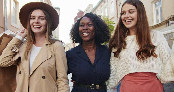 Happy pretty multi-ethnic diverse females walking in town in sunlight. Portrait of beautiful smiling women holding shopping bags. African American and Caucasian girls talking outdoor. Emotions concept
