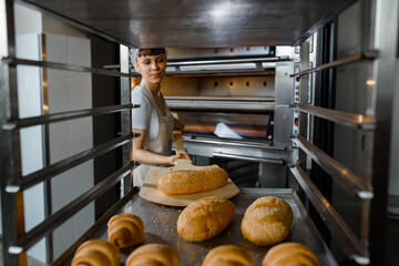 Close up of young caucasian woman baker putting the fresh bread on the shelves/rack at baking manufacture factory.