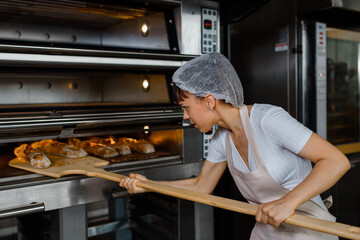Young caucasian woman baker is taking off from oven the french baguette bread with wood peel at...