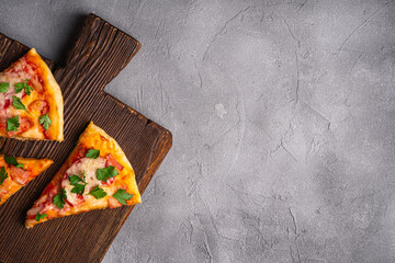 Hot pizza slices with mozzarella cheese, ham, tomato and parsley on brown wooden cutting board, stone concrete background, top view copy space for text