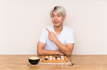 Young asian man with sushi in a table pointing finger to the side