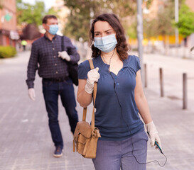 Focused woman wearing medical mask and rubber gloves listening to music on earphones on way to work along city street on spring day. New life reality during COVID 19 pandemic