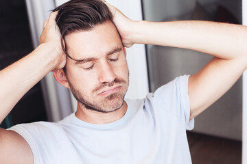 A close up portrait of a young casual man in a t-shirt looks disappointed and stressed, eyes closed, holding his head  