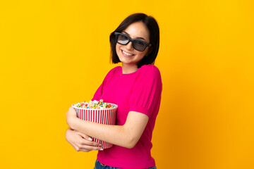 Fototapeta premium Young caucasian woman isolated on yellow background with 3d glasses and holding a big bucket of popcorns