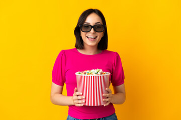 Young caucasian woman isolated on yellow background with 3d glasses and holding a big bucket of popcorns