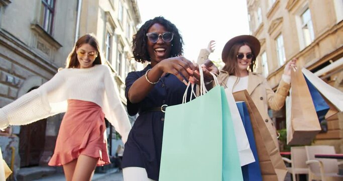 Cheerful mixed-races girls in sunglasses with shopping bags walking on street. Happy Caucasian beautiful girls moving in city. African American satisfied female after shopping outdoor. Fun concept
