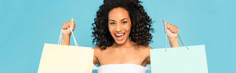 panoramic shot of excited african american woman holding shopping bags and smiling isolated on blue