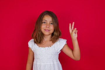 Little caucasian girl with blue eyes wearing white dress standing over isolated red background showing and pointing up with fingers number two while smiling confident and happy.