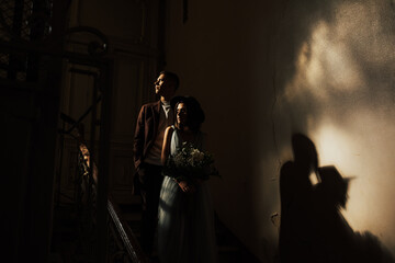 Newlyweds in love stand in an old dark building near the bright window and embrace. Portrait of a stylish groom with coat and a beautiful cute bride in a blue dress and black hat. Wedding day.