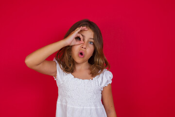 Little caucasian girl with blue eyes wearing white dress standing over isolated red background doing ok gesture shocked with surprised face, eye looking through fingers. Unbelieving expression.