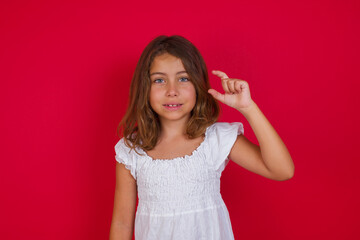 Little caucasian girl with blue eyes wearing white dress standing over isolated red smiling and confident gesturing with hand doing small size sign with fingers looking and the camera. Measure concept