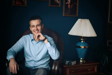 man with blue eyes in a blue shirt sitting in a chair smiling, propping his chin with his hand,...