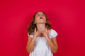 young brunette kid standing against red background amazed and surprised looking up and pointing with fingers and raised arms.