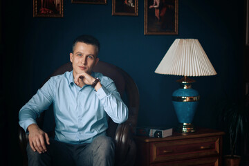 man with blue eyes in a blue shirt sitting in a chair smiling, propping his chin with his hand,...