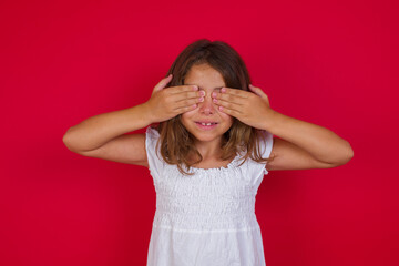 Little caucasian girl with blue eyes wearing white dress standing over isolated red background covering eyes with hands smiling cheerful and funny. Blind concept.