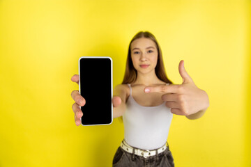 Naklejka premium Pointing on phone's screen. Caucasian young girl's portrait isolated on yellow studio background. Beautiful female blonde model. Concept of human emotions, facial expression, sales, ad, youth culture