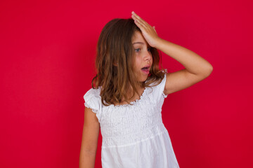Little caucasian girl with blue eyes wearing white dress standing over isolated red background surprised with hand on head for mistake, remember error. Forgot, bad memory concept.