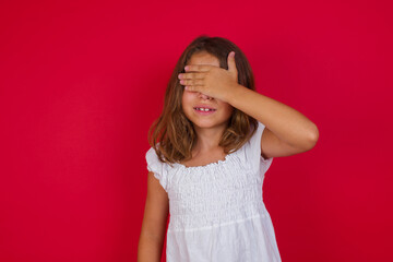 Little caucasian girl with blue eyes wearing white dress standing over isolated red background smiling and laughing with hand on face covering eyes for surprise. Blind concept.