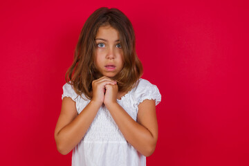 Little caucasian girl with blue eyes wearing white dress standing over isolated red background praying for luck has hands crossed near face, amazed and opening mouth looking front.