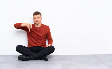 Young handsome man sitting on the floor showing thumb down sign