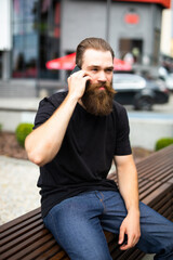 Handsome young bearded man wearing jacket sitting on a bench and talking on mobile phone while looking at documents