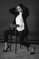 dark-haired girl straightens her hair while sitting on a chair, turned back forward in a black suit, white shirt and black high-heeled shoes