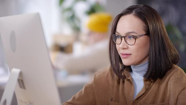 Tilt up shot of beautiful young Asian woman in glasses sitting at her desk in office and working on computer. Color swatches lying on table