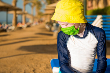 sad boy in green mask sitting on sunbed on empty beach line 