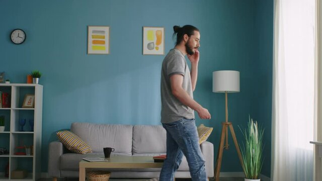 Young man is walking around the living room, talking on the phone, discussing daily tasks with his colleague, busy and motivated, Slow motion.