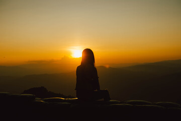Silhouette of a girl sitting on a cliff side looking at the sunset.  Beautiful sunset background.