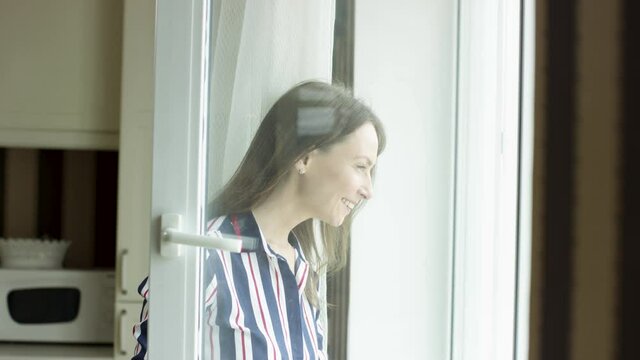 Dark haired smiling woman opens the door of a balcony and looks outside. Video. Side view of a happy lady at home looking outdoors through a window of a living room on a sunny day.