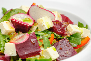 Vegetable salad with radish, soft cheese, beetroot and greens in a white plate on a gray background. Snacks on the restaurant.