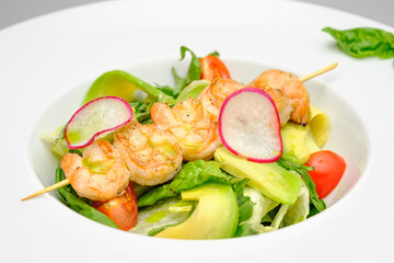 Grilled shrimp on a skewer with vegetables on a white plate on a gray background.