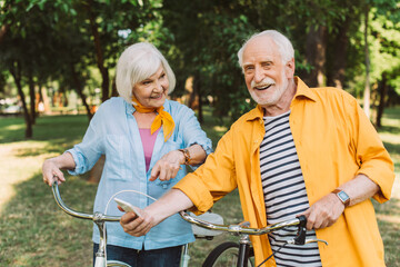 Smiling elderly woman pointing with finger near husband with smartphone and bikes in park