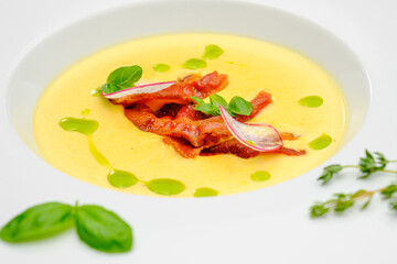 Corn cream soup with smoked meat in a white plate decorated with basil, on a gray background. The first course on the restaurant menu.